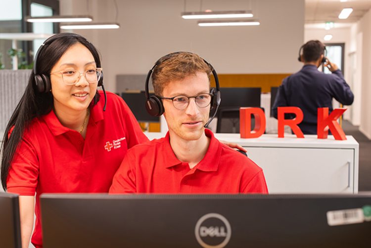 Dieses Bild zeigt zwei Mitarbeitende des DRK in DRK-Poloshirts mit Headsets auf dem Kopf, die gemeinsam auf einen Monitor gucken.