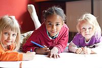 Dieses Imagebild zeigt drei Kinder, die auf dem Boden liegen, malen und direkt in die Kamera gucken.
