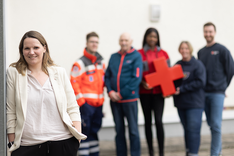 Dieses Imagebild zeigt eine diverse Gruppe an Menschen, die teilweise DRK-Kleidung tragen. Eine Frau lehnt im Vordergrund gegen eine Wand. Die restlichen Personen stehen im Hintergrund.