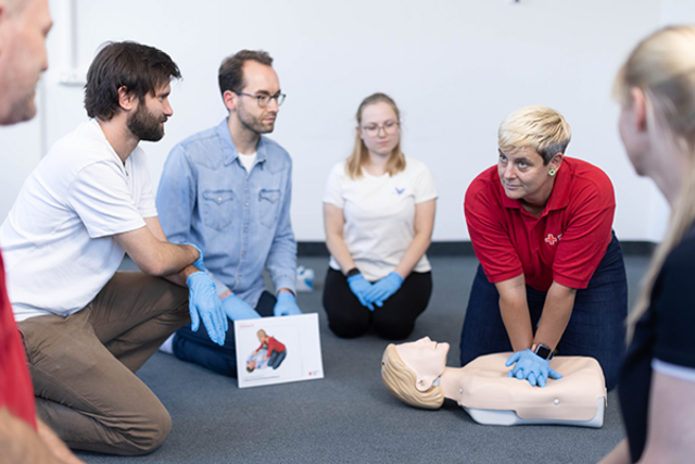 Dieses Bild zeigt eine Schulungssituation einer Rotkreuzkurses für Erste Hilfe. Die Kursleiterin demonstriert den teilnehmenden die Herzdruckmassage an einer Übungspuppe.